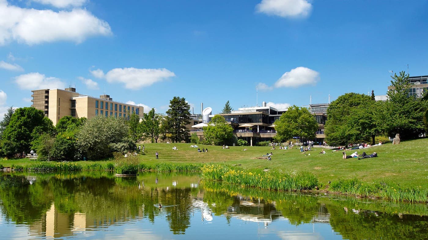 Claverton Down kampüsündeki göl, çimenlikte oturan öğrenciler ve arkadaki ders binaları, University of Bath