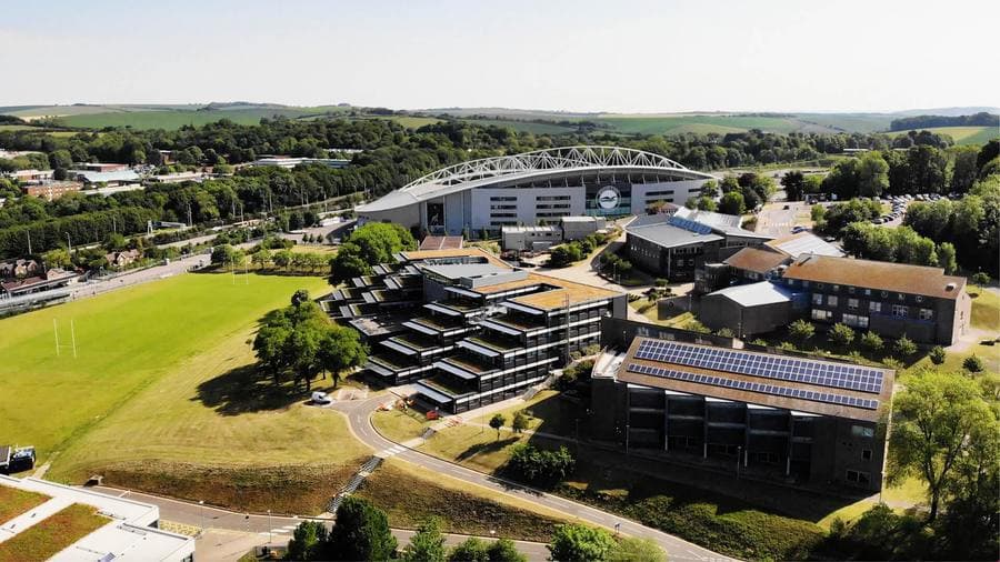 Falmer kampüsünün havadan görünümü; arka planda Falmer Stadyumu ve South Downs tepeleri, Brighton Üniversitesi